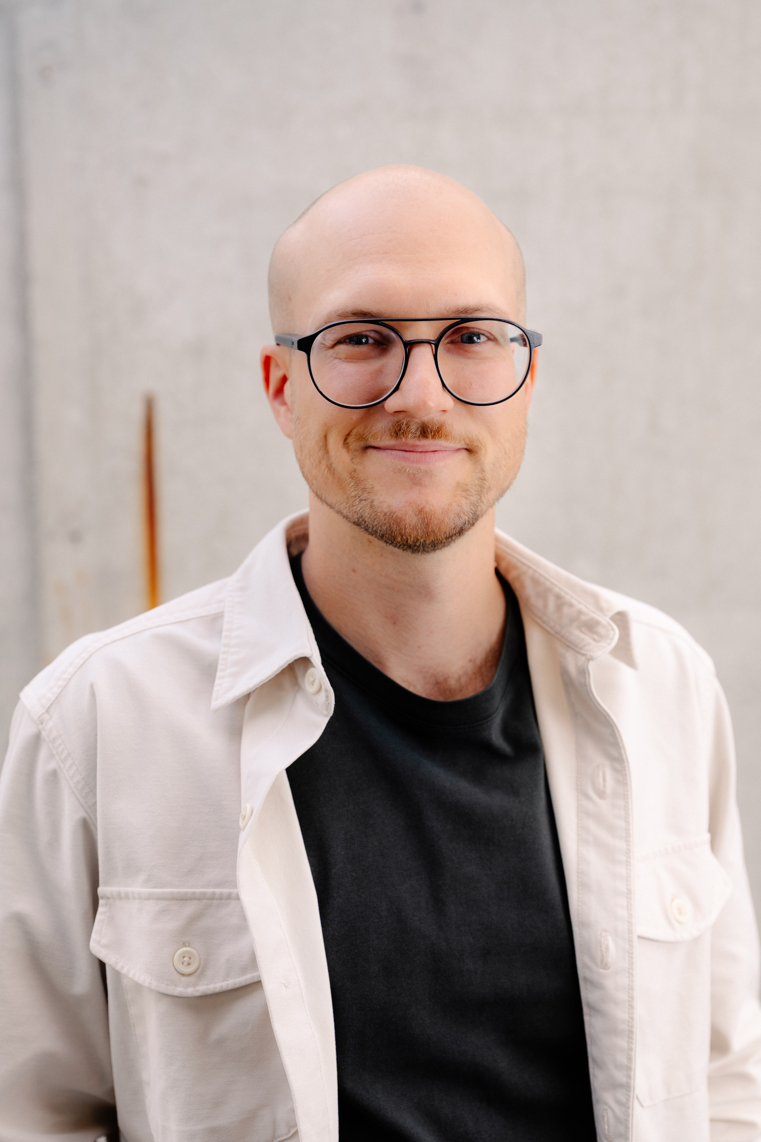 L&auml;chelnder Mann mit Brille und wei&szlig;er Jacke vor Betonwand